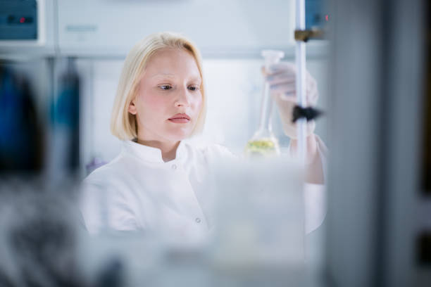 Blonde female scientist wearing a lab coat, working in her laboratory and conducting diverse experiments using scientific tools at her disposal, carefully holding a Volumetric flask in her hands and examining the liquid inside it