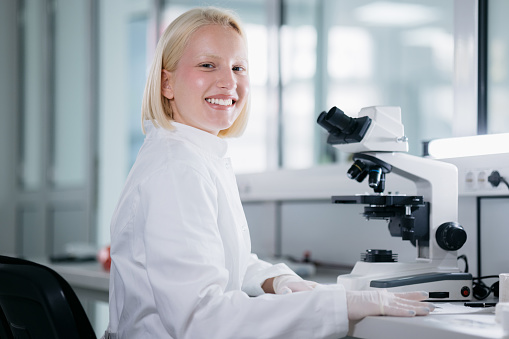 Portrait of a happy blonde female scientist wearing a lab coat, working in her laboratory and conducting diverse experiments, using a microscope  and looking at the camera