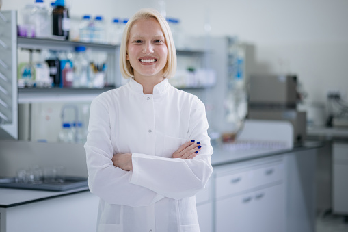 Portrait of a confident blonde female scientist wearing a lab coat and standing in her laboratory next to her equipment, looking at the camera with her arms crossed