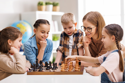 Happy female teacher in glasses pointing at board and explaining moves to cheerful children during chess lesson in daytime at school