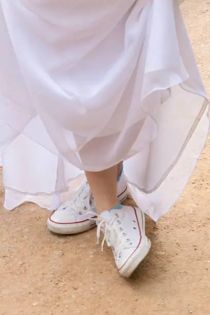 Woman in white dress and white tennis shoes holds up the bottom of dress to show off tennis shoes. The woman is standing on dirt