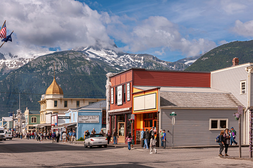 Skagway, Alaska, USA - July 20, 2011: Retail stores on Broadway with Golden North Hotel tower in back under blue cloudscape and snow covered mountain in back. Facades, clothing, advertisements add colors