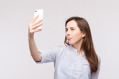 Studio portrait of pretty young Caucasian girl with long brunette hair in casual striped shirt making funny face and sticking tongue at camera while making selfie on cell phone..