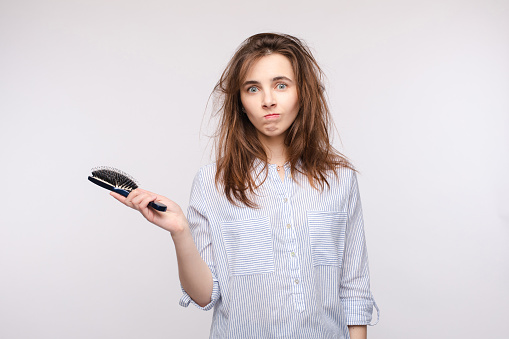 Studio portrait of young adult woman with messy brunette hair holding a hairbrush in right hand and looking at camera with puzzlement and confusion. Isolate on white.