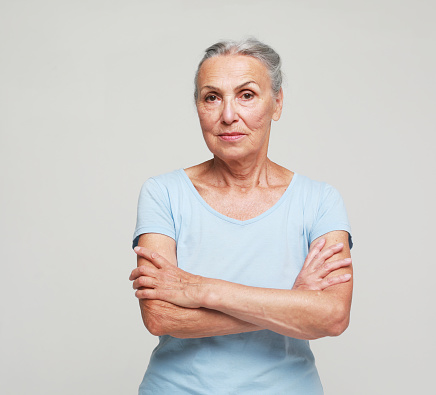 Mature elderly woman standing over grey background. Old senior female, 60s gray-haired lady looking at camera arms crossed, portrait close up.