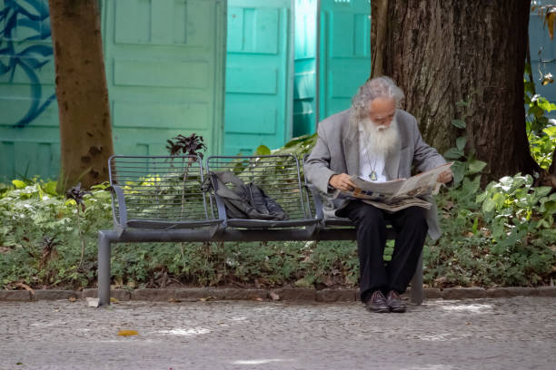 JUIZ DE FORA, BRAZIL - JUNE 05, 2022 - Elderly man sitting on a bench at Parque Halfed square in downtown Juiz de Fora, Brazil on a Sunday morning and reading a newspaper.