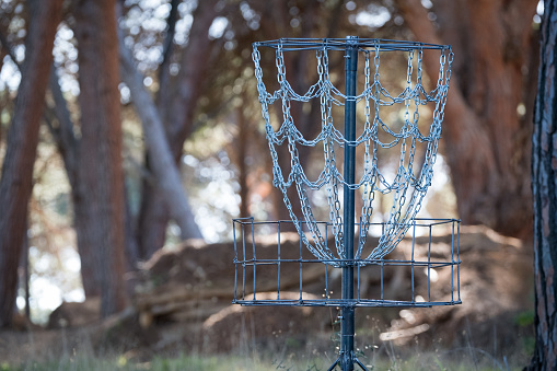 A Disc Golf Basket stands in a beautiful forest. Disk golf is a sport where one throws various disks through a challenging course into a basket.