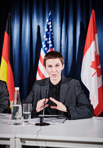 Portrait of young woman with short hair speaking in microphone at table during press conference with flags in background