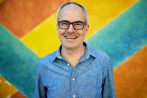 Close-up of Caucasian man with short gray hair and eyeglasses in blue long sleeved shirt standing against colorful background and smiling at camera.