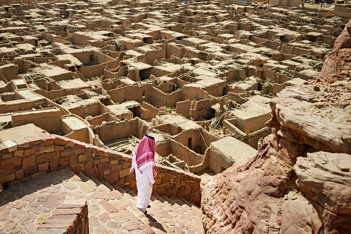 Elevated view of Middle Eastern man in dish dash, kaffiyeh, and agal walking down staircase with ruins of Al-Ula mudbrick houses in background.