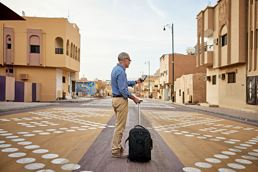 Three-quarter rear view of tourist in casual attire pulling wheeled luggage and pausing to check portable information device for directions in Al-Ula.