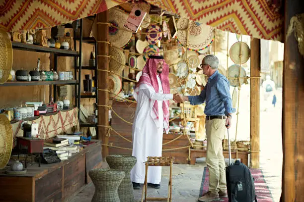 Mature Caucasian professional with wheeled luggage greeting young Middle Eastern man in traditional attire during trip to develop new commercial alliances.