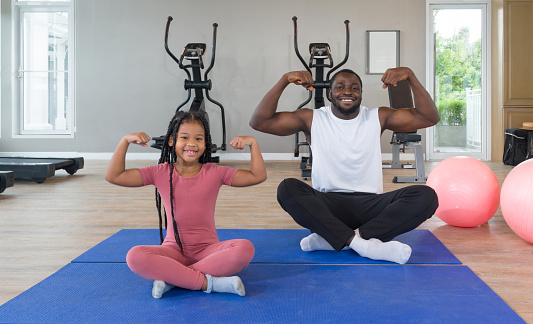 Black Cornrow Braids girl in sportswear flexing muscles, biceps, triceps with her father on yoga mat. Two yoga ball are placed next to them. Happy family enjoy holiday together in f