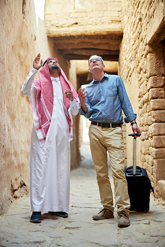 Full length view of young Middle Eastern man explaining cultural heritage to business traveler with luggage as they walk through mudbrick alleyway.