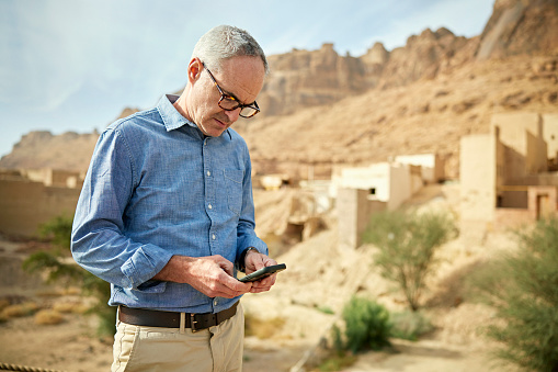 Three-quarter front view of Caucasian man in casual attire standing outdoors and checking portable information device with historic district in background.