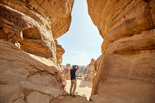 Full length rear view of mature Caucasian man standing within Jar Rock aka Jabal Jarrah and photographing surrounding rock formations in Al-Ula Valley.
