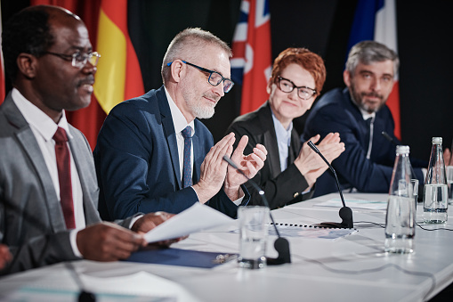 Upset displeased senior politician in suit standing up and talking about his position loudly at meeting, government representatives attentively listening to him