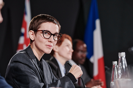 Portrait of young female speaker in eyeglasses looking at camera while speaking in microphone at conference