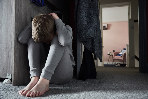 A young boy sat on the floor of his bedroom with his head in his hands feeling sad