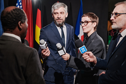 Group of people, male politician confronted by journalists with microphones.