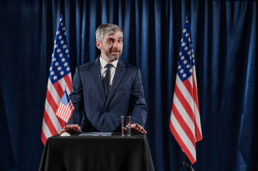 Friendly middle aged politician in suit gesticulating and talking with audience from podium against USA flag during election debate