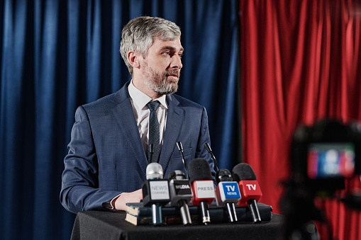 Mature bearded businessman standing at stage with microphones and giving an interview during online broadcast at press conference