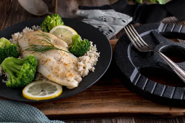 Homemade fresh cooked fitness meal for sports nutrition and muscle gain with a  seared fish fillet, brown rice and broccoli sprouts. Served with dumbbell on a rustic and wooden table background. Closeup view.
