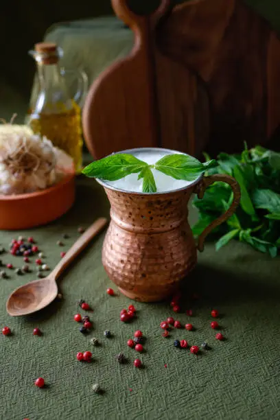 Istanbul, Turkey-May 31, 2022: Homemade buttermilk with fresh mint leaves in copper mug on khaki green tablecloth. Traditional Turkish drink "Ayran".    There are chili pepper balls scattered on the tablecloth. Behind mug are wooden cutting boards, a bunch of fresh mint, a bottle of olive oil and garlic. Iced ayran. Shot with Canon EOS R5.