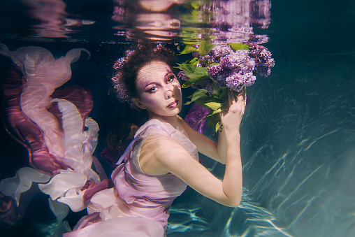 woman with lilac bouquet in colorful clothes on the dark background swimming underwater