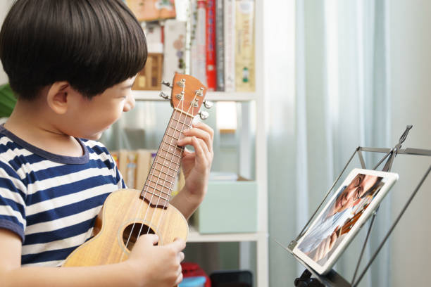 istock Happy and cute Asian elementary age boy playing ukulele and videocalling with teacher via computer tablet 1401690030