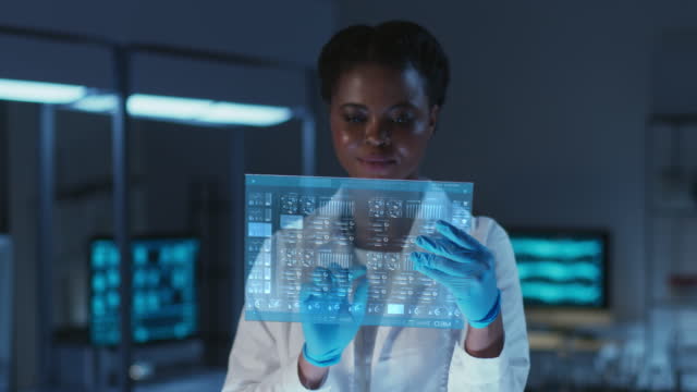Beautiful young African- American scientist in a lab coat and protective gloves working on futuristic HUD screen in a modern laboratory. Scientist with HUD in a dark blue color scheme