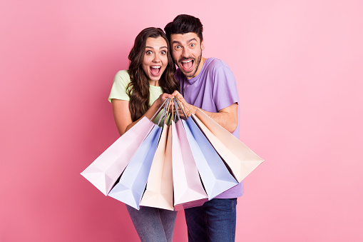 Portrait of attractive trendy amazed cheerful couple carrying new things hugging black friday isolated on pink pastel color background.