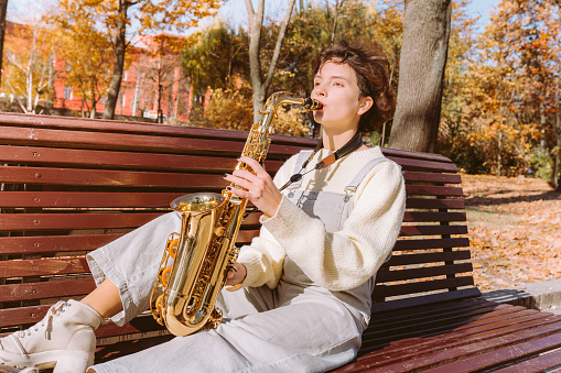young beautiful young lady musician sits alone on bench in autumn park, plays melody on saxophone