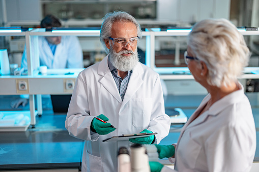 A Caucasian senior scientist with a colleague discussing the experiment results. Male and female professional researchers wearing lab coats, working and collaborating in a laboratory.