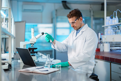 A young adult Caucasian biochemist in his 30s wearing a lab coat and protective glasses. He is mixing liquid chemicals using a pipette in a modern lab.