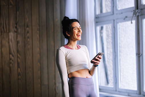 A muscular Caucasian female in sports clothes smiling while watching something on her smartphone after doing her workout at home.