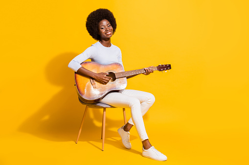 Full size profile portrait of pretty positive dark skin lady sit playing guitar isolated on yellow color background.