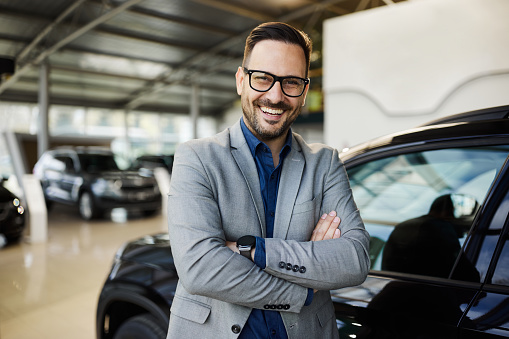 Happy male salesperson standing in a car showroom with crossed arms and looking at camera.