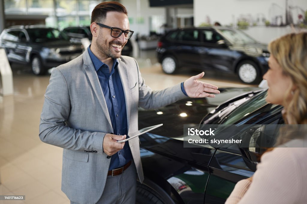 Happy salesman selling the car to his female customer in a showroom. Happy car salesman talking to his customer while showing her a car in a showroom. Car Dealership Stock Photo
