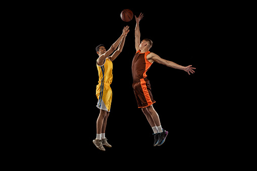 Dynamic portrait of two young men, professional basketball players in a jump, throwing ball into basket isolated over black studio background. Concept of sport, team game, action, active lifestyle, ad
