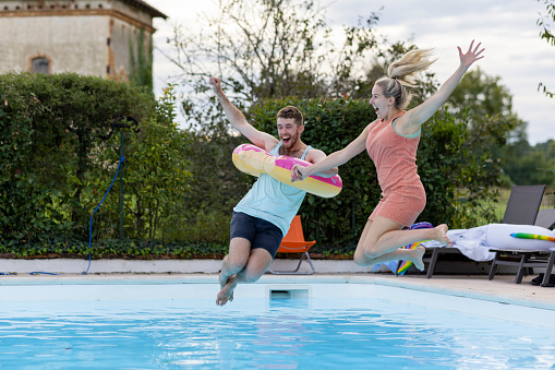 A medium wide angle view of two friends holding hands and smiling as they jump into a swimming pool whilst enjoying a trip away to Toulouse in the South of France.