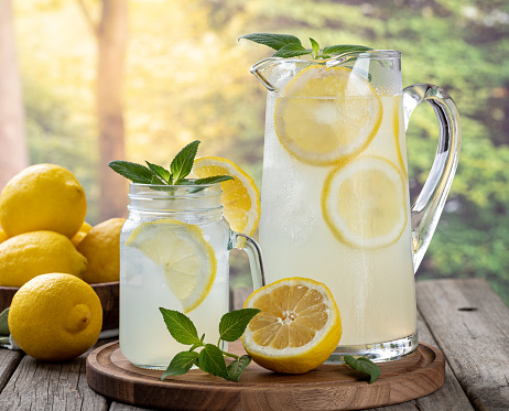 Glass and pitcher of lemonade with lemon slices and mint on an old wooden table with rural summer background