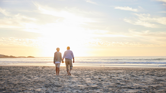 Rear view of a senior couple walking hand in hand toward the ocean surf at sunset during a trip to the beach