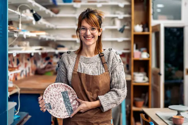 Stock photo of smiley woman wearing apron holding ceramic piece, smiling and looking at camera.