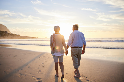 Rear view of an affectionate senior couple walking hand in hand along a sandy beach at sunset in summer
