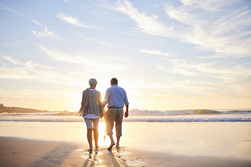Rear view of a loving senior couple walking hand in hand together along a sandy beach at sunset in the summertime