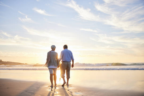 kochająca para seniorów spacerująca ręka w rękę po plaży o zachodzie słońca - men footprint beach sunset zdjęcia i obrazy z banku zdjęć