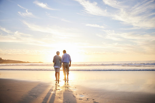 Rear view of an affectionate senior couple walking hand in hand together along a sandy beach at sunset in the summertime