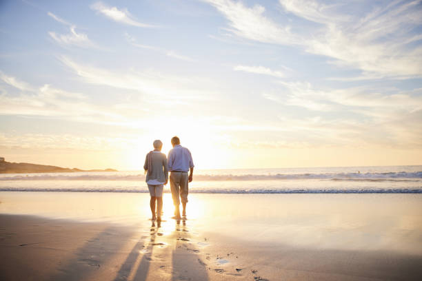 kochająca para seniorów spacerująca ręka w rękę po piaszczystej plaży o zachodzie słońca - men footprint beach sunset zdjęcia i obrazy z banku zdjęć
