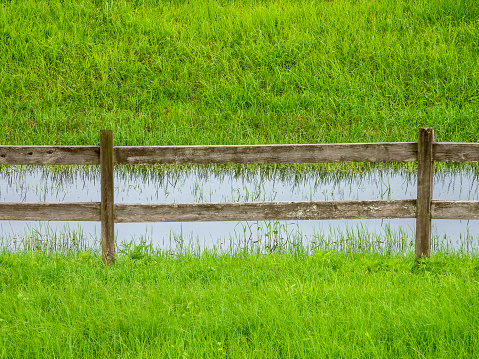 Rail fence at the edge of a ditch full of stormwater at the bottom of a grassy hill in a conservation area, once a landfill, on a cloudy summer afternoon in southwest Florida